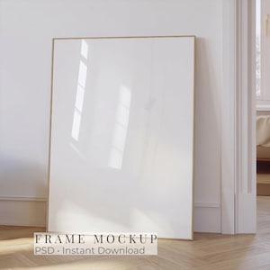Puede incluir: Un marco de fotos rectangular vertical con un borde fino de madera clara y un centro blanco vacío. El marco presenta una superficie brillante con reflejos de luz visibles. El texto "FRAME MOCKUP PSD - Instant Download" está impreso abajo.