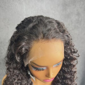 Peut inclure: Perruque bouclée brun foncé présentée sur une tête de mannequin. La perruque a un devant en dentelle et une racine des cheveux d'apparence naturelle. Les boucles sont serrées et tombent sur les côtés. Le mannequin a un teint neutre, avec des cils peints et des lèvres roses.