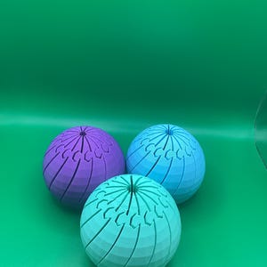 May include: Three spherical objects with a segmented, interlocking design. One sphere is purple, one is light blue, and one is teal. Each sphere features a series of vertical, curved grooves that meet at a central point at the top and bottom.