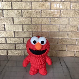 Pode incluir: Um personagem de brinquedo de pelúcia vermelho com olhos brancos grandes, um nariz laranja brilhante e um amplo sorriso preto. O brinquedo tem uma aparência texturizada e difusa e está em pé. O fundo apresenta uma parede de tijolos dourados.