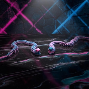 May include: Two articulated snake toys in shades of blue and pink are displayed on a reflective surface. The snakes have a textured, segmented design. The background features neon pink and blue lighting, creating a vibrant and dynamic scene.