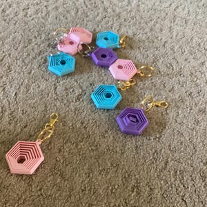 Op de afbeelding: Een verzameling kleurrijke geometrische sleutelhangers in roze, blauw en paars. Elke sleutelhanger heeft een gelaagd zeshoekig ontwerp en een goudkleurige sleutelring. De sleutelhangers liggen verspreid op een neutraal gekleurd tapijt.