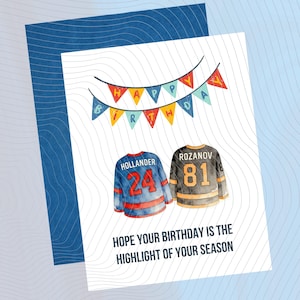 Puede incluir: Tarjeta de cumpleaños con sobre azul. La tarjeta presenta ilustraciones de acuarela de dos camisetas de hockey, una azul y roja con el número 24, y la otra negra y dorada con el número 81. Una pancarta dice "Happy Birthday". El texto dice "Espero que tu cumpleaños sea lo más destacado de tu temporada."