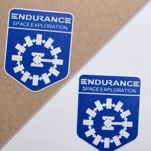 May include: Two blue and white stickers with the text "ENDURANCE SPACE EXPLORATION". Each sticker features a gear-like design with a central emblem. The stickers are on a white and brown surface.