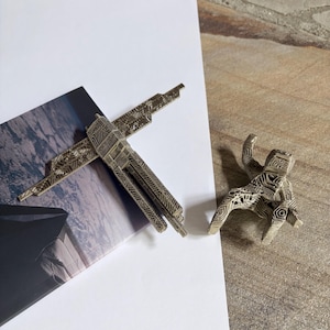 May include: Two intricately designed, metallic sculptures. One resembles a cross, the other a stylized human figure. Both have detailed patterns and are set against a white background and a textured surface.