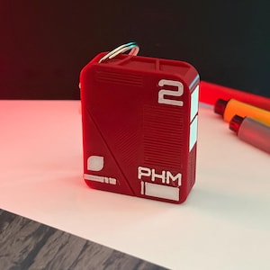 May include: A red rectangular keychain with the number "2" and the letters "PHM" in white. The keychain has a silver key ring attached. The background is a gradient of black and red.