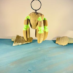 May include: A hanging keychain featuring a central tan, textured, rounded body with four articulated, segmented limbs. The limbs alternate between tan, wood-grain textured sections and bright neon green joints. It hangs from a black metal ring.