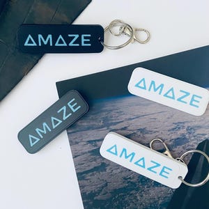 May include: Rectangular keychains with the word "AMAZE" in a light blue font. The keychains are black, gray, and white. Each keychain has a silver keyring and clasp.