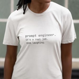 以下が含まれることがあります： 「prompt engineer. it's a real job. stop laughing.」という黒い文字がプリントされた白いTシャツ。半袖で丸首です。