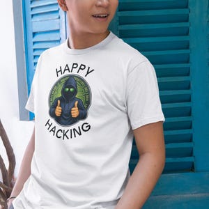 Peut inclure: T-shirt blanc avec un motif d'un personnage encapuchonné aux yeux verts brillants faisant un signe de pouce, entouré de code binaire. Les mots "HAPPY HACKING" sont imprimés au-dessus et en dessous du motif dans un design circulaire.