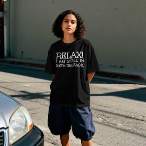 Könnte beinhalten: Schwarzes T-Shirt mit dem weißen Text "RELAX! I AM STILL IN BETA RELEASE." Die Person trägt eine marineblaue Shorts. Die Person steht auf einer Straße neben einem Auto.