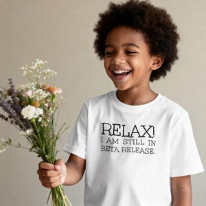 May include: A child wearing a white t-shirt with the text "RELAX! I AM STILL IN BETA RELEASE." smiles broadly. They hold a colourful bouquet of flowers. The background is a plain, neutral tone.