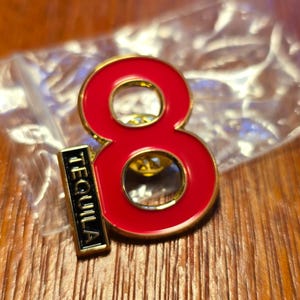 Puede incluir: Pin de esmalte rojo con forma del número ocho con borde dorado. Un pequeño rectángulo negro con la palabra "TEQUILA" en letras blancas está adjunto. El pin está sobre una superficie de madera.