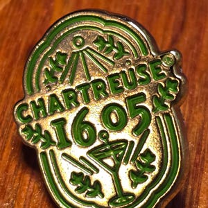 Puede incluir: Pin dorado con borde verde y texto verde "CHARTREUSE 16.05". El pin presenta una copa de cóctel y hojas decorativas. Es probable que el pin sea un artículo de colección o material promocional.