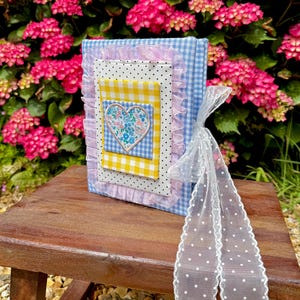 May include: A rectangular fabric-covered album featuring a light blue and white gingham pattern. The front is decorated with a white polka-dot panel, a yellow gingham square, and a floral heart appliqué, all framed by a ruffled pink trim.