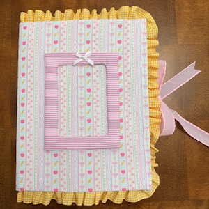 May include: A fabric-covered photo album featuring a light blue background with vertical stripes of pink bows, red apples, and yellow carrots. A pink and white striped rectangular frame with a small white bow sits in the center. The album has a yellow gingham ruffle border and a pink ribbon tie.