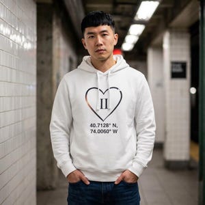 May include: White hoodie with a black heart design containing the Roman numeral II. Below the heart is the text "40.7128° N, 74.0060° W". The hoodie has a drawstring hood and a front pocket.