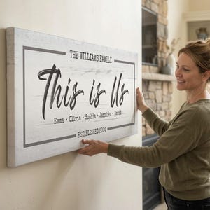以下が含まれることがあります： 黒い筆記体で「This is Us」と書かれた長方形の白い木製サイン。サインには、家族の名前、家族の名前、そして「Established 2004」という年号も含まれています。そのサインは人が持っています。
