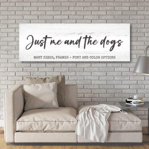 May include: A rectangular white wooden sign with the text "Just me and the dogs" in black script. Below the text are the words "MANY SIZES, FRAMES + FONT AND COLOR OPTIONS". The sign is hanging on a white brick wall above a beige sofa.