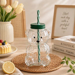 May include: A clear glass bear-shaped drinking cup with a green lid and a green and white striped straw. The cup sits on a woven mat. A lemon half and a small cake are in the foreground.