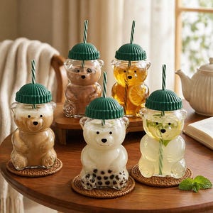 May include: A collection of bear-shaped glass drinkware with green lids and straws. The glasses are filled with various beverages, including coffee, tea, and a drink with boba. The glasses sit on small woven coasters.
