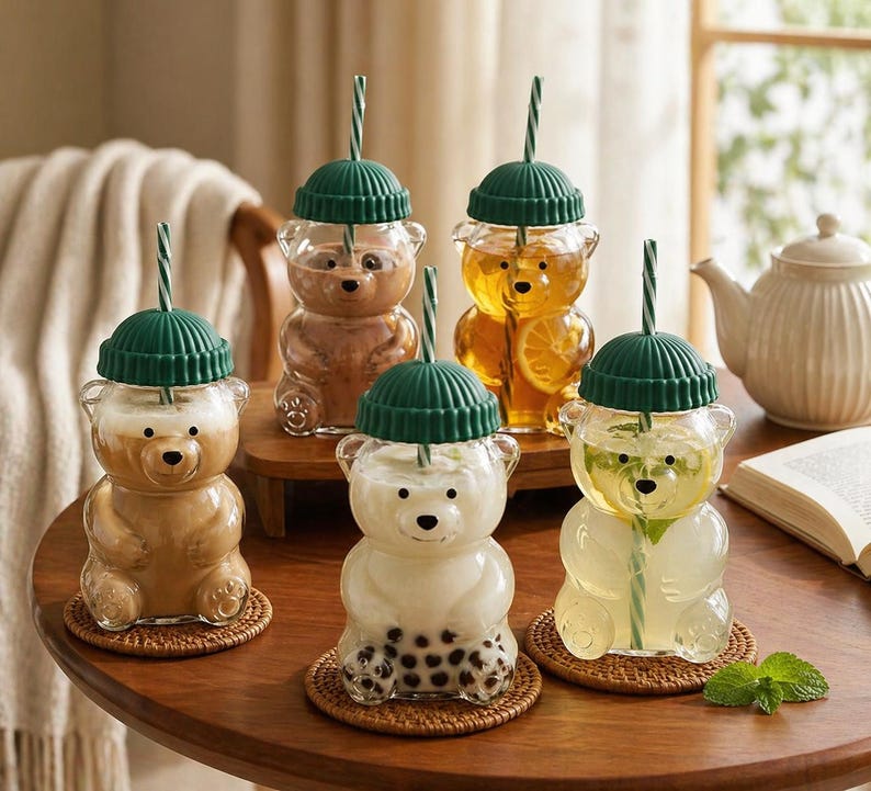 May include: A collection of bear-shaped glass drinkware with green lids and straws. The glasses are filled with various beverages, including coffee, tea, and a drink with boba. The glasses sit on small woven coasters.