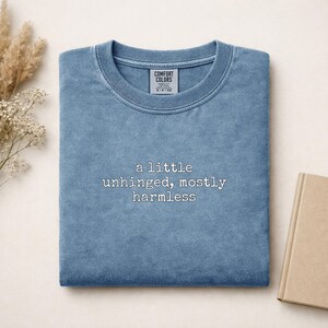 Könnte beinhalten: Ein blaues T-Shirt mit Rundhalsausschnitt und Vintage-Waschung. Das Kleidungsstück zeigt mittig einen weißen Stickschriftzug mit dem Text: a little unhinged, mostly harmless. Ein Etikett der Marke Comfort Colors ist im Kragen sichtbar.