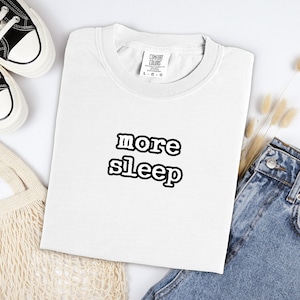 Könnte beinhalten: Ein weißes Kurzarm-T-Shirt mit dem Text more sleep in schwarzer, fetter, umrandeter Schrift, mittig auf der Brust platziert. Das T-Shirt hat einen Rundhalsausschnitt und ein Comfort Colors Markenetikett im inneren Kragenbereich.
