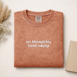 Può includere: Una t-shirt a girocollo color terracotta con il testo ricamato in bianco "no thoughts, head empty" centrato sul petto. La maglietta presenta un'etichetta del marchio Comfort Colors all'interno del colletto e uno scollo a coste.