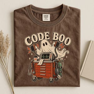 May include: A brown crew neck t-shirt featuring a graphic of a ghost holding defibrillator paddles above a medical crash cart. The text CODE BOO is printed in a curved font above the ghost. The cart includes a monitor, stethoscope, and oxygen tank.