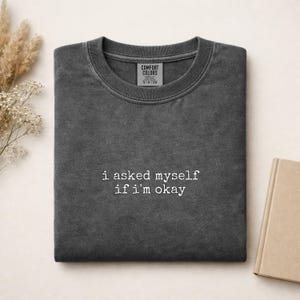 Könnte beinhalten: Ein anthrazitfarbenes T-Shirt mit Rundhalsausschnitt und Vintage-Waschung. Das Shirt zeigt den gestickten weißen Text "i asked myself if i'm okay" mittig auf der Brust. Ein Markenetikett von Comfort Colors ist im Halsausschnitt sichtbar.