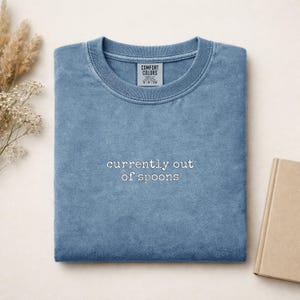 May include: A blue crewneck t-shirt with a vintage wash finish. The centre of the chest features white embroidered text that reads "currently out of spoons". The shirt has a Comfort Colors brand label visible inside the collar area.