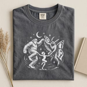May include: A charcoal gray t-shirt featuring a white graphic print of a Bigfoot, a moth-like creature, and an alien holding hands in a circle under a crescent moon and stars. The inner neck label displays the text Comfort Colors.