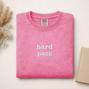 May include: A bright pink crewneck t-shirt with a ribbed collar and the text "hard pass" embroidered in white on the center chest. The interior neck label reads "Comfort Colors" with size information visible on the garment tag.
