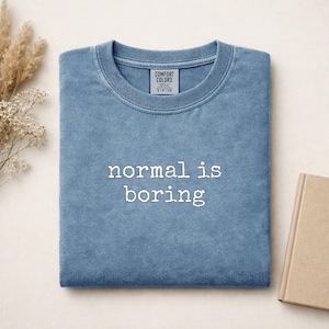 Może przedstawiać: Niebieska bluza z okrągłym dekoltem i wykończeniem typu vintage-wash. Napis "normal is boring" jest wyhaftowany białą nicią na środku klatki piersiowej. Wewnętrzna metka na karku zawiera napis "COMFORT COLORS" oraz rozmiar S-P-CH pod nazwą marki.