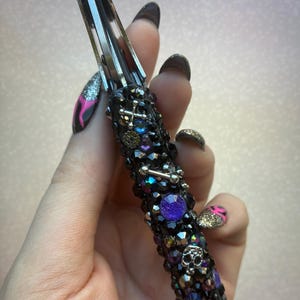 May include: A decorative, embellished pocket knife with a black handle encrusted with black and iridescent beads, crosses, and a skull. The knife's blade is silver. A large purple gem is set in the handle. The knife is held in a hand with long, dark nails.