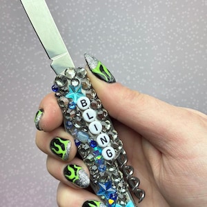 Könnte beinhalten: Ein dekoratives Taschenmesser mit einer silbernen Klinge und einem Griff, der mit Strasssteinen und blauen Akzenten verziert ist. Das Wort "BLING" ist auf dem Griff geschrieben. Am Fuß des Griffs befindet sich eine kleine blaue Schleife.