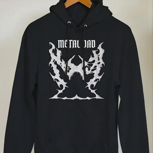 Könnte beinhalten: Schwarzer Kapuzenpullover mit dem Text "METAL DAD" in einer gotischen Schriftart. Das Design zeigt eine weiße Grafik eines Schädels und Flammen. Der Kapuzenpullover hängt an einem Holzbügel.