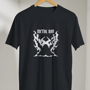 Könnte beinhalten: Schwarzes T-Shirt mit dem Text "METAL DAD" in weißer, gotischer Schrift über einer weißen Grafik. Die Grafik ist ein stilisiertes, flammenartiges Design. Das T-Shirt hängt an einem Holzbügel.