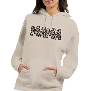 Può includere: Una felpa con cappuccio beige con tasca a marsupio e cappuccio con coulisse. La parola "MAMA" è stampata sul petto con un motivo leopardato nero e marrone. La felpa è realizzata in materiale morbido.