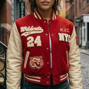 Op de afbeelding: Rode en crèmekleurige varsity-jas met het woord "Wildcats" en "Est. 1928" in witte letters. De jas heeft het nummer "24", een tijgerpatch en de letters "M.Y.C. NY". De mouwen zijn crèmekleurig.