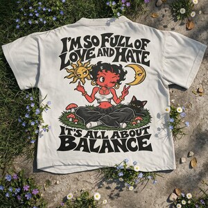 Könnte beinhalten: Ein cremefarbenes T-Shirt mit einer Grafik einer rot häutigen Figur, die in einem grasbewachsenen Feld mit einer schwarzen Katze, einer Sonne und einem Mond sitzt. Der Text lautet, I'M SO FULL OF LOVE AND HATE IT'S ALL ABOUT BALANCE, umgeben von kleinen roten Pilzen.