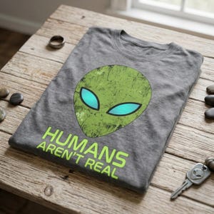 Könnte beinhalten: Ein graues T-Shirt mit einem abgenutzten grünen Alien-Kopf-Motiv und leuchtend blauen Augen. Unter dem Alien-Kopf ist der Text "HUMANS AREN'T REAL" in einer fetten, lindgrünen serifenlosen Schrift gedruckt. Das Shirt hat einen Rundhalsausschnitt.