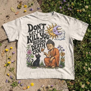 May include: A cream-coloured short-sleeve t-shirt featuring a graphic print with the text "DON'T LET LIFE KILL YOU BEFORE DEATH CAN". The design includes a seated Buddha figure, a black cat, a butterfly, flowers, and a sun and moon illustration.