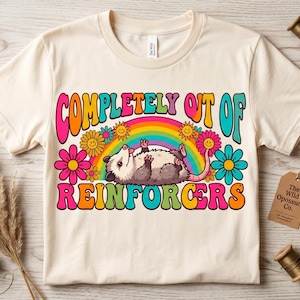 May include: Cream-colored t-shirt featuring a cartoon opossum lying on its back under a rainbow. The shirt has the text "COMPLETELY OUT OF REINFORCERS" in colorful, retro-style lettering, surrounded by flowers.