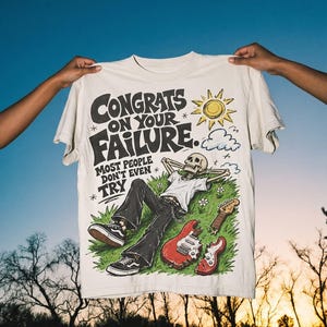 May include: Off-white t-shirt with the text "Congrats on Your Failure" and a graphic of a skeleton relaxing on grass with guitars. The design includes a sun, clouds, and flowers. The shirt is held up against a blue sky.