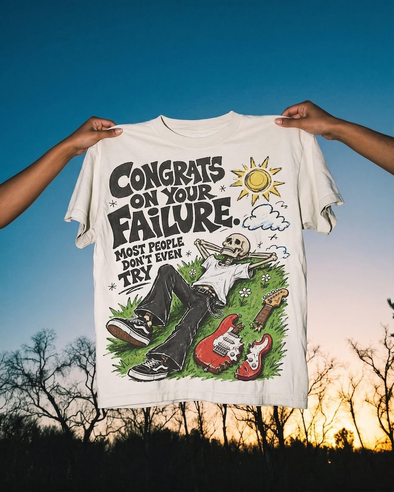 May include: Off-white t-shirt with the text "Congrats on Your Failure" and a graphic of a skeleton relaxing on grass with guitars. The design includes a sun, clouds, and flowers. The shirt is held up against a blue sky.