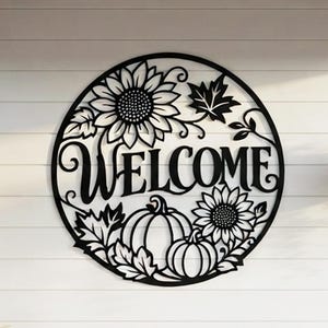 May include: A circular black metal wall sign featuring the word WELCOME in a script font, surrounded by cut-out designs of two sunflowers, two pumpkins, and several maple leaves, creating a decorative seasonal display for an outdoor space.