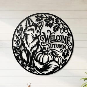 Puede incluir: Una pieza de arte mural circular de metal negro con un diseño recortado de un zorro sentado junto a calabazas, tallos de trigo y hojas de roble. El texto "WELCOME AUTUMN" está integrado en el diseño con una tipografía decorativa.