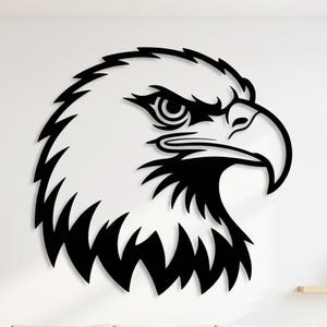 Op de afbeelding: Een zwart metalen wandkunstwerk dat de kop van een adelaar afbeeldt. De kop van de adelaar is in profiel weergegeven, met gedetailleerde kenmerken zoals het oog, de snavel en de veren. Het kunstwerk is bevestigd aan een witte muur boven een beige bank.